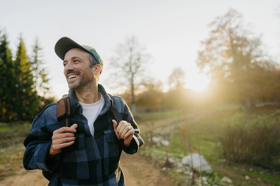 Smiling man in a plaid flannel jacket and cap carries a backpack while hiking outdoors on a wooded trail at sunset.